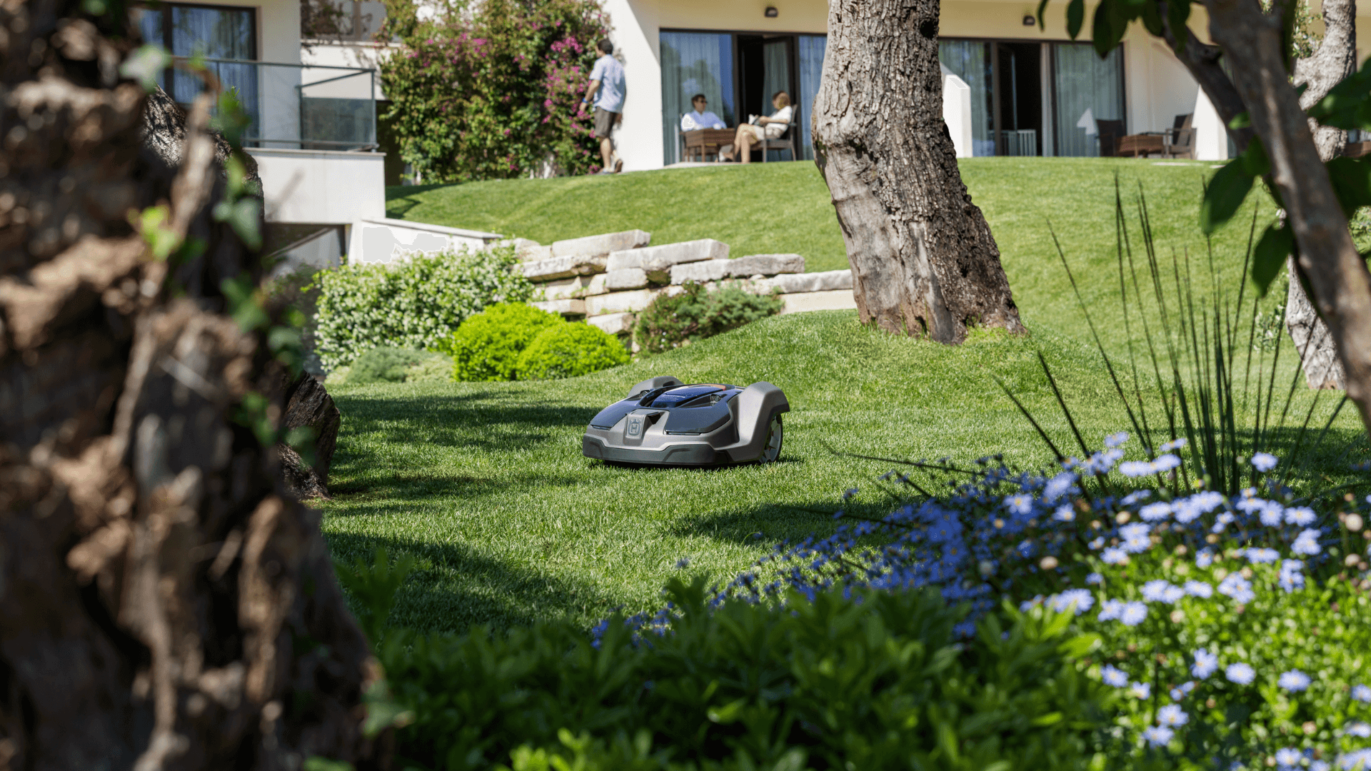 Automower sitting in grass patch in backyard with family cooking bbq in background